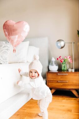 Baby standing and holding on the the side of a bed with a heart balloon