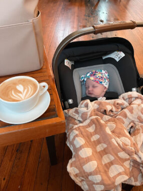Baby sleeping in a car seat on the floor next to a table with a bag and coffee